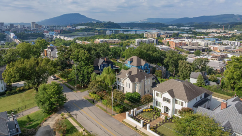 Aerial view of a suburban neighborhood with elegant homes, tree-lined streets, and a distant city skyline. A bridge spans a river, set against rolling hills and a cloudy sky.