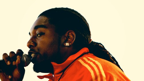 Rapper Traxamillion holds a microphone close to his mouth and wears a bright orange track jacket.