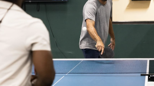 Two people playing table tennis. We can't see their faces because the image is too low, and the other has their back turned, but they are stood by a blue table, with the ball in play hovering over the net.