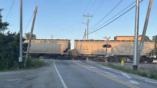 A freight train crosses a rural road at sunset. The crossing gate is down, lights flashing. Utility poles lean nearby, creating a calm and still atmosphere.