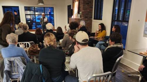 A packed audience at the Kenwick Table look on as a speaker stands at the microphone reading from a piece of paper.