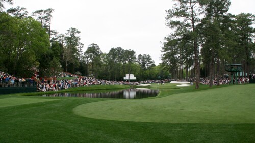 A view of a green gold course with spectators lining the opposite side of a pond in its middle.