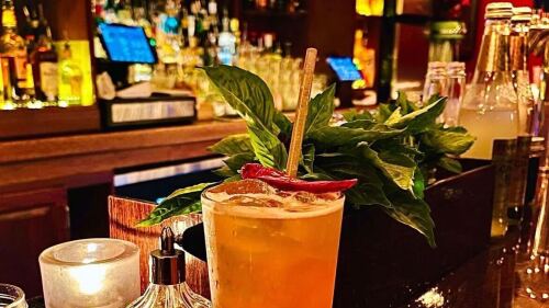 In a dimly lit bar, a bright orange cocktail with a chili pepper, a straw, and a green plant stand in the foreground.