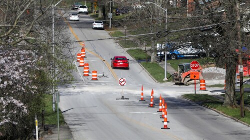 An intersection with cones going down the middle and detour signs, construction machinery, and piles of rocks.