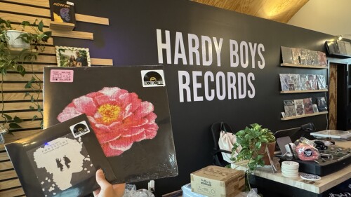 Two records being held in front of a counter and Hardy Boys Records wall.