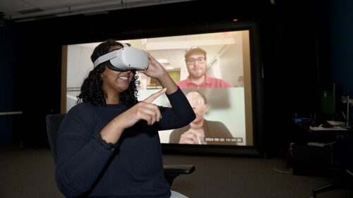 A student wear virtual reality goggles with a screen of a Zoom call in the background.