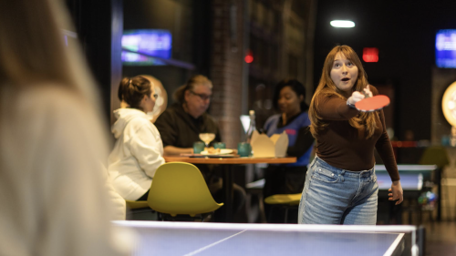 People playing table tennis in a bar. The table is blue, the person hitting the ball is smiling, and there are people sat a table behind them speaking to a server, possibly ordering food.