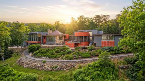 Modern house with colorful geometric facade and large windows, surrounded by lush greenery and trees. A serene, sunlit sky is in the background.