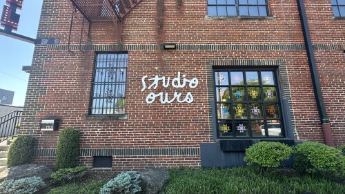 Red brick building with large black-framed windows, adorned with colorful star decorations. "Studio Ours" is painted in white letters. Lush green bushes and sidewalk in the foreground.