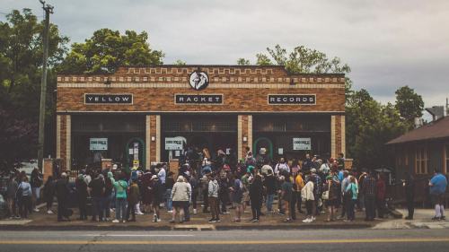 Crowd gathered outside a brick building named "Yellow Racket Records," with people chatting and waiting in line. Overcast sky, trees in the background.