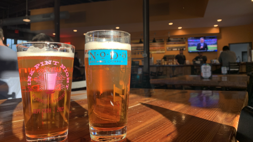 Two beers on a table with a taproom in the background. 