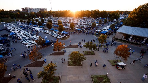 A bird's eye view of a parking lot filled with cars, canopies, and fans tailgating.