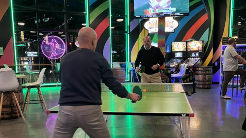 Two people playing table tennis in a neon-lit room. There are pinball machines behind them, and the two are wearing business-casual attire. It is dark outside, seen through large floor-to-ceiling windows.
