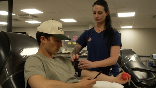 A nurse prepares to draw blood from a seated man holding a stress ball. The room is brightly lit, and the atmosphere is calm and professional.