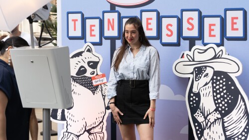 Woman smiling in front of a backdrop reading "Greetings from Tennessee." Cartoon raccoon and armadillo with cowboy hats flank her, creating a playful vibe.