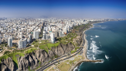 An aerial shot of the coastline that lines Lima Peru. A vast number of buildings line the land atop cliffs, with a deep blue ocean on the right of the image, running alongside a road beneath the cliffs.