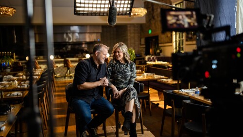 A couple sitting in the middle of a restaurant dining room with a camera pointed at them. 