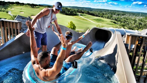 An adult sits in the back of a double tube and a kid sits in the front, both with their hands in the air, as w lifeguard begins pushing the tube down a water slide.