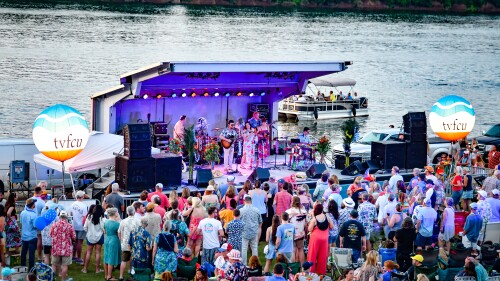 Outdoor concert by a river with a vibrant crowd in colorful summer clothing. A band performs on stage under bright lights, with boats visible in the water.