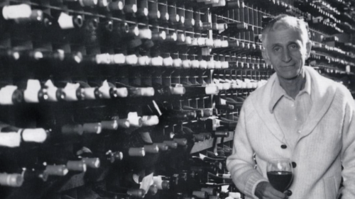 Bern Laxer, founder of Bern's, stands in front of his wine collection in a black and white photo. He is holding a glass of red, and wearing a light colored woolen cardigan, looking at the camera with a wry smile.