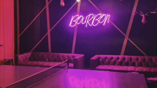 A table tennis tale is in the foreground, with a purple neon sign that says 'BOURBON' on the wall behind it. There are two long leather couches, and fake deer heads on the wall.