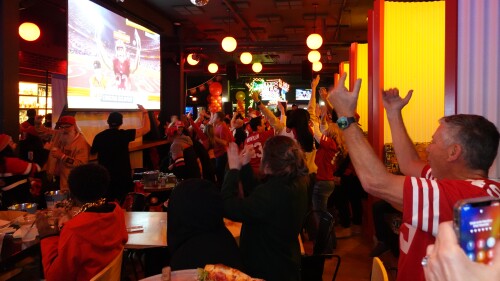 A large group of people wearing mostly red in a dimly lit event space cheer on the 49ers game playing on a large screen projector.