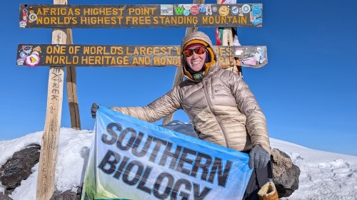A person in winter gear smiles while holding a "Southern Biology" banner at the snowy Uhuru Peak, Mount Kilimanjaro. Colorful summit signs are visible.