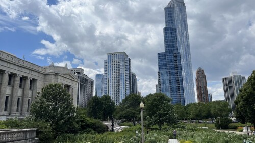 Urban skyline with modern glass skyscrapers under a partly cloudy sky. In the foreground, a lush garden and historic building create a serene contrast.
