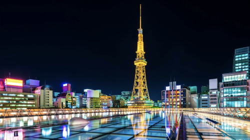 Illuminated Nagoya TV Tower glowing gold against a dark night sky, surrounded by modern city buildings, with colorful neon lights reflecting across a glass rooftop in the foreground.