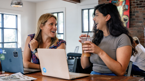 Two City Editors drinking coffee and smiling with their laptops in front of them. 