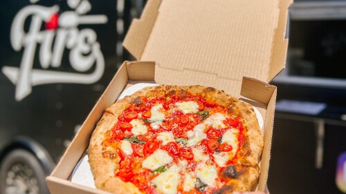 A woman's hand holding an open box of pizza in front of a black food truck. 