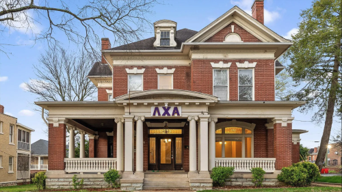 Large brick fraternity house with tall columns, a wide porch, and Greek letters on top. The building has white trim and a welcoming foyer.