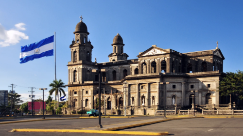 Historic stone cathedral with two bell towers under a clear blue sky. A large Nicaraguan flag flutters on the left, evoking a sense of national pride.