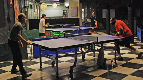 People playing table tennis in a room with black and white checkered floor. There are people either side of two tables, competing under lights. The tables are blue, and the participants look focused on the games at hand.