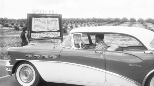 A man sits inside a car as a woman reads a sign about the legend of Spook Hill