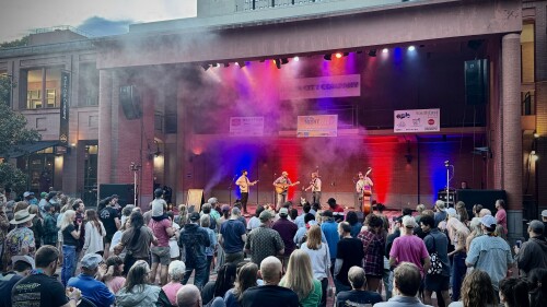 Outdoor concert with a lively crowd watching a band perform on a lit stage. Colorful lights and smoke create a vibrant, energetic atmosphere.