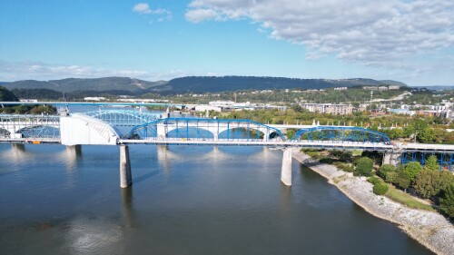 Aerial view of a blue truss bridge spanning a wide river. Green trees and a rocky shoreline are visible, with distant hills under a partly cloudy sky.