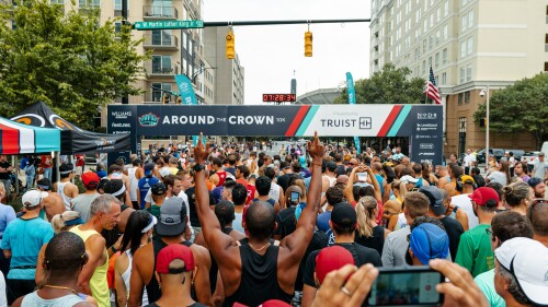 A group of runners with a person pointing to an Around the Crown 10k banner in Uptown Charlotte.
