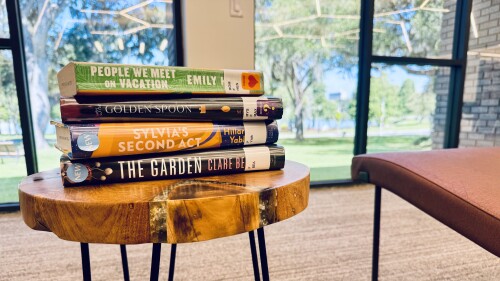 A stack of books is shown on a table in Lakeland Public Library with windows looking out at Lake Morton.