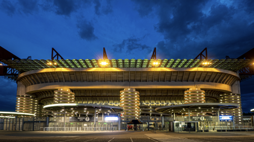 A photograph of the iconic San Siro Stadium in Milan, lit up at night. The sky is cloudy and dark blue overhead, with the stadium lit up by a flurry of lights on its concourses. Empty entrance gates are in front of the giant structure.