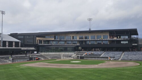 A newly constructed baseball stadium under a cloudy sky, featuring empty stands and a well-maintained field. The modern building in the background has glass windows.
