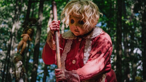 A person in a creepy doll mask with blond curls, wearing a red, vintage dress, holds a stick in a forest. Dolls hang eerily from trees, enhancing the unsettling atmosphere.