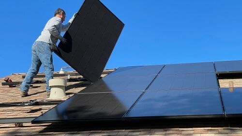 Someone in the middle of laying a solar panel down on a roof next to already installed panels.