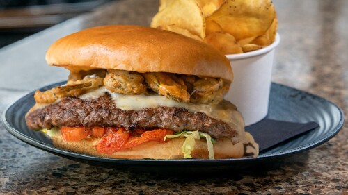 The Fireworks Burger from Harry's is shown with ghost pepper cheese, fried pickled jalapeños, and lettuce on a bun. Chips in a cup are behind the burger.