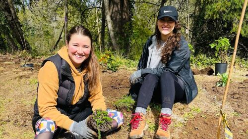 Two people wearing tights, jackets, hiking shoes, and gloves sit on ground recently disturbed by planting activity on a sunny day. 