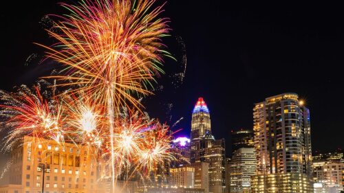 Fireworks exploding over a baseball field with people sitting on the field and the Charlotte skyline in the background.