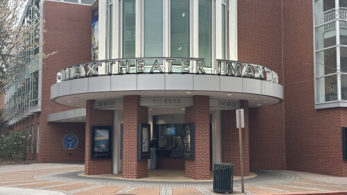 Exterior of a brick IMAX theater with a modern design, featuring a tall, glass-front entrance. The sign reads "IMAX THEATER." Atmosphere is calm.
