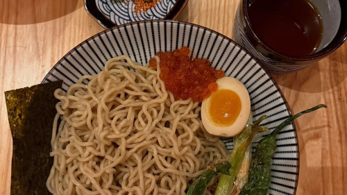 A bowl filled with ramen noodles, a boiled egg, roe, nori, and bok choy, with a cup of tea and a cup of roe on the side.