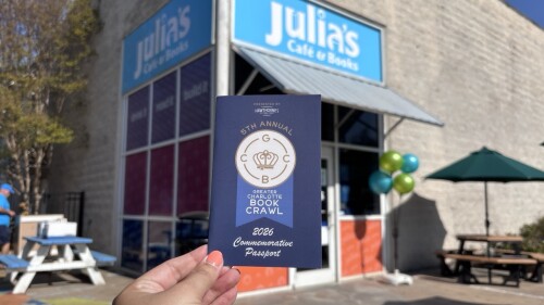 A hand holding a small blue Book Crawl passport in front of a bookstore.