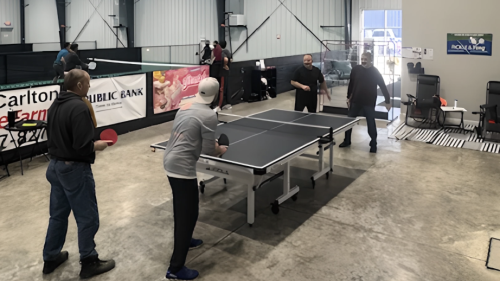 A group of people playing table tennis in a warehouse. There are two people either side of a table, with pickleball being played in the background.